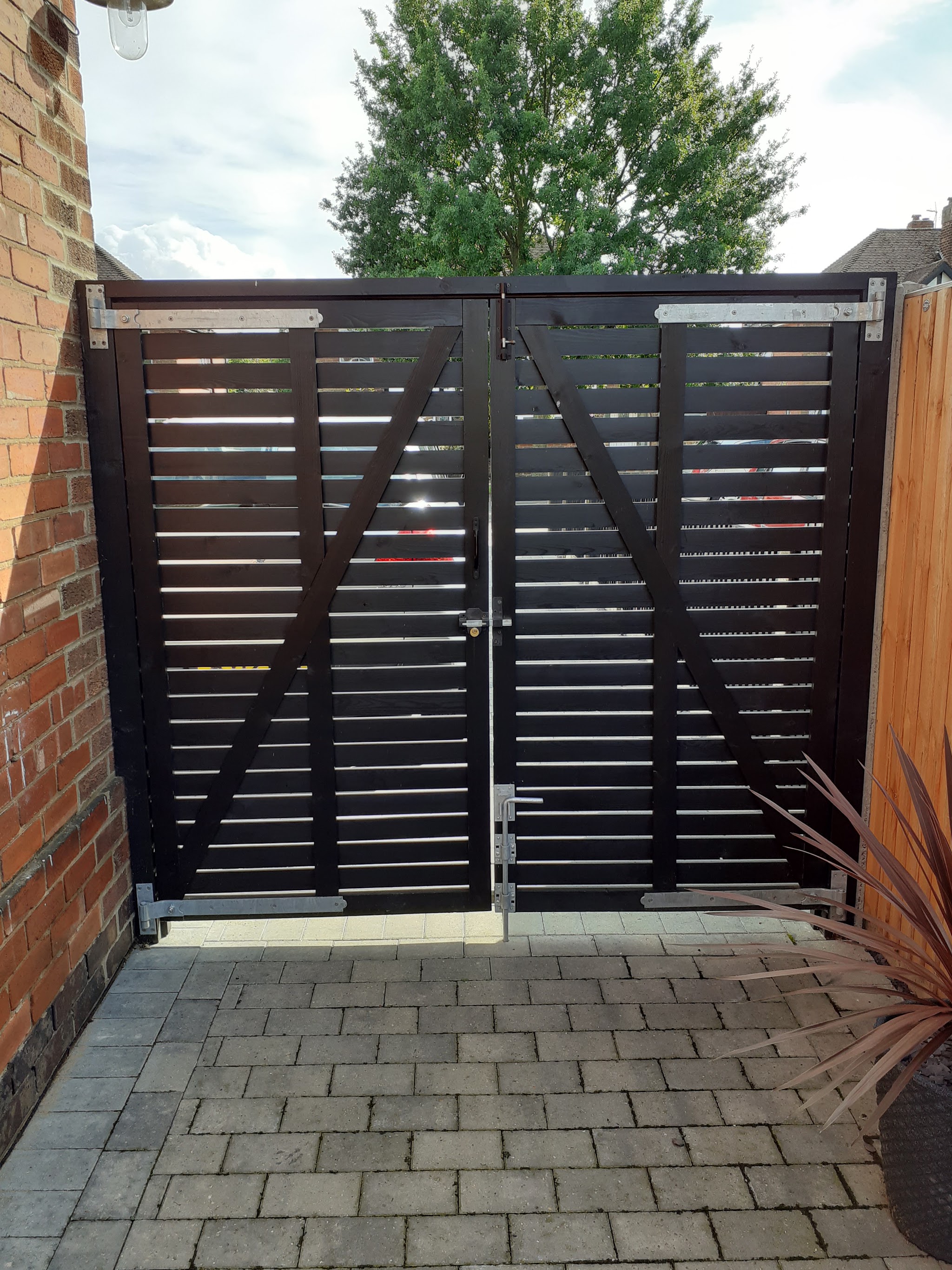Black slatted timber double gates with galvanised hinges over a block-paved drive
